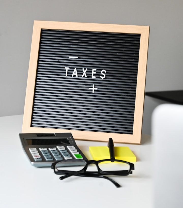 Calculator, glasses, and a yellow sticky note on a desk next to a framed 'TAXES' sign.